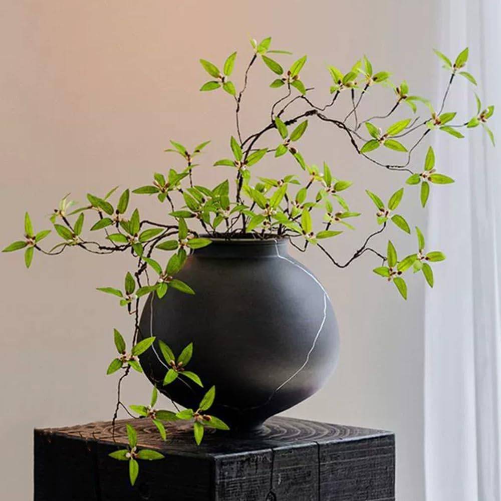 Faux greenery stems arranged in a dark ceramic vase on a wood console beside soft window light, styled for calm neutral home decor.