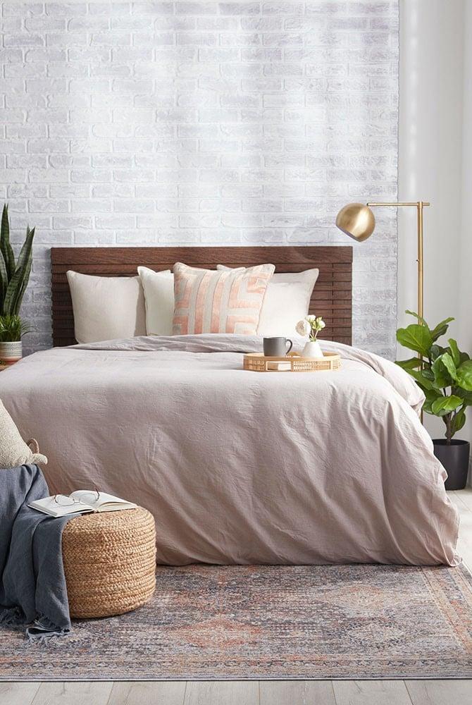 Cozy neutral bedroom with a wood bed, blush bedding, patterned area rug, gold floor lamp, woven pouf, potted plants, and bright natural light.