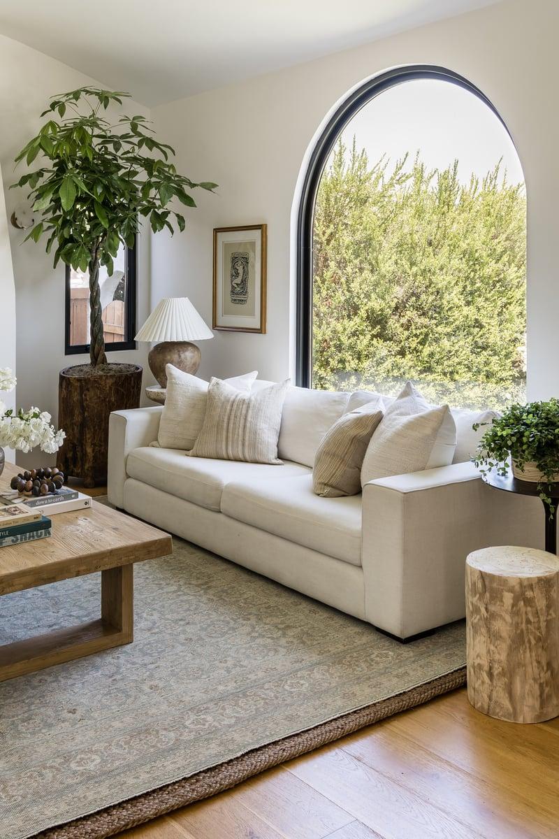 Bright neutral living room with a white couch, arched window, layered rug, wood coffee table, books, lamp, and leafy plants in warm natural light.