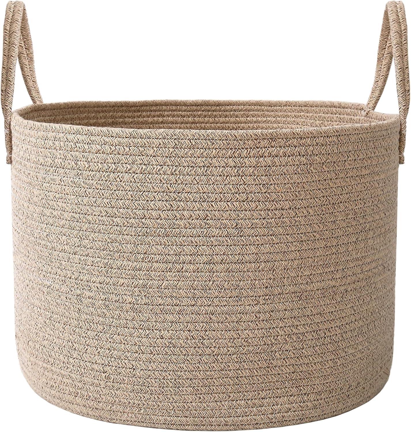 Large oatmeal woven rope basket with handles styled beside a neutral sofa, filled with soft blankets and pillows in a bright warm living room.