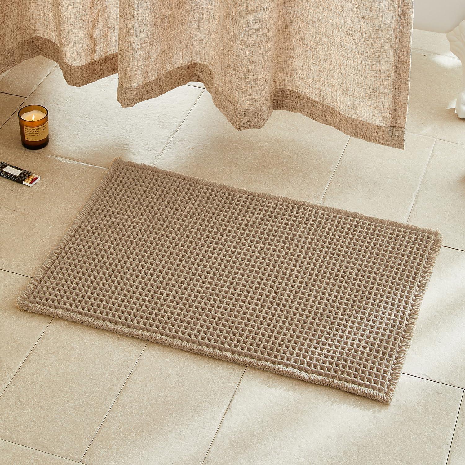 Khaki waffle bath mat on beige tile beside a soft linen shower curtain, amber candle, and warm natural light for cozy neutral bathroom decor.